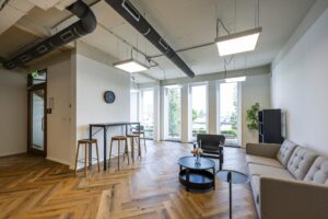 Moderne lounge- en wachtruimte aan de De Ruijterkade met grote ramen, houten visgraatvloer en stijlvolle zitmeubels.